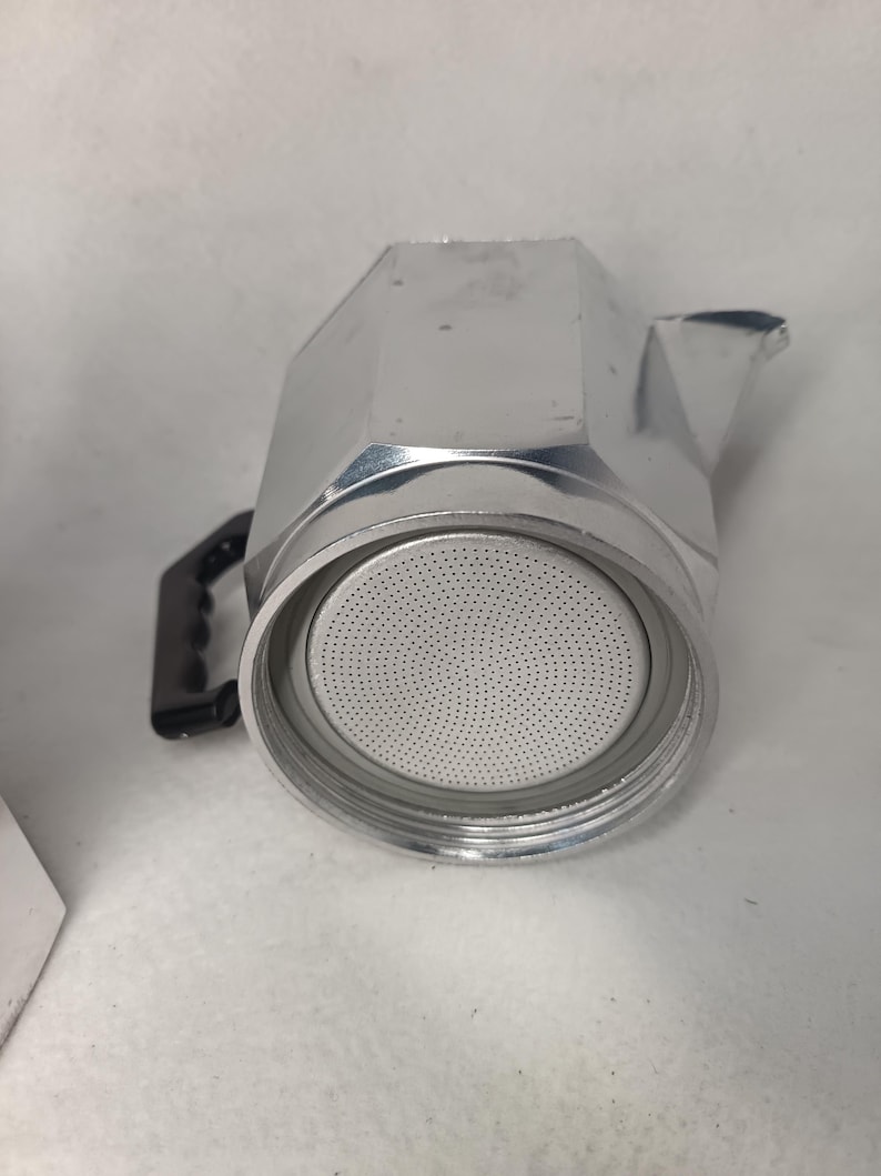 May include: A silver stovetop espresso maker with a black handle. The coffee pot is made of metal and has a round filter basket. The spout is on the right side of the pot. The coffee maker is designed to brew espresso.