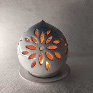 May include: A white ceramic essential oil diffuser with a floral design. The diffuser has leaf-shaped cutouts that emit a warm orange glow. It sits on a matching white saucer, creating a soft, inviting ambiance.
