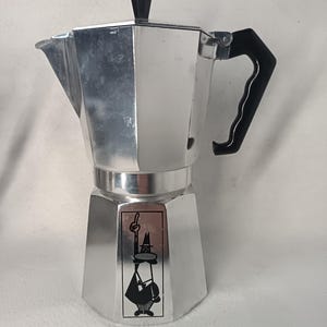 May include: A silver stovetop espresso maker with a black handle and lid knob. The coffee pot has a distinctive octagonal shape and a black and white cartoon figure on the front.