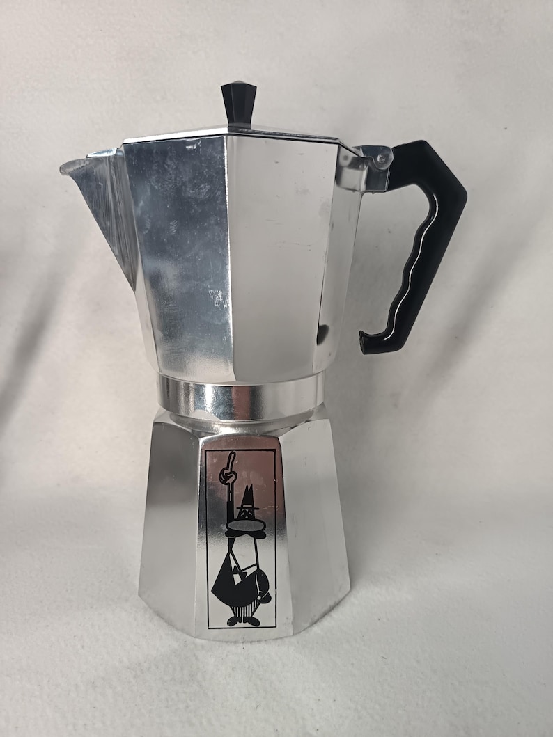 May include: A silver stovetop espresso maker with a black handle and lid knob. The coffee pot has a distinctive octagonal shape and a black and white cartoon figure on the front.