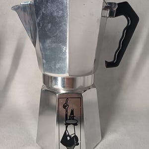 May include: A silver stovetop espresso maker with a black handle and lid. The coffee pot has a geometric design and a black and white graphic on the front. This Italian-style coffee maker is designed for brewing espresso.