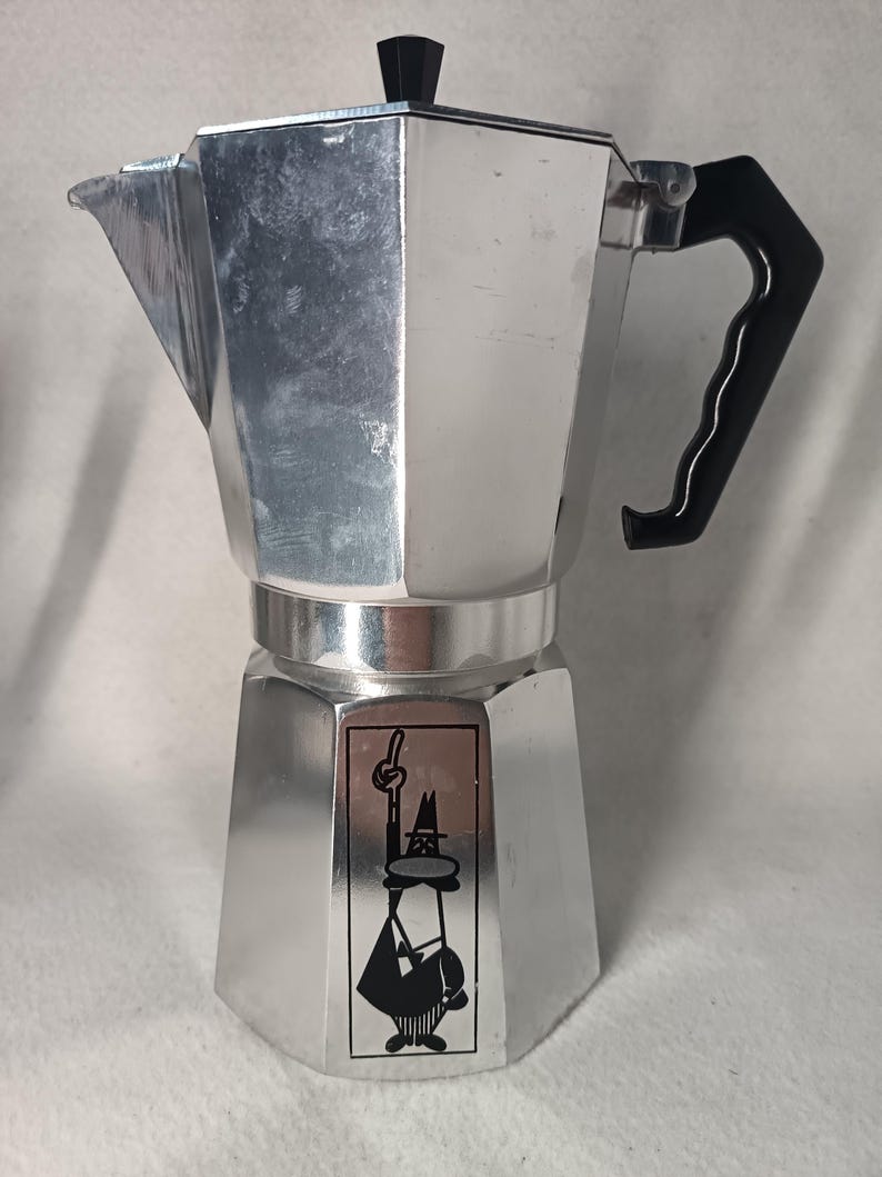 May include: A silver stovetop espresso maker with a black handle and lid. The coffee pot has a geometric design and a black and white graphic on the front. This Italian-style coffee maker is designed for brewing espresso.