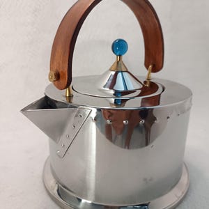 Könnte beinhalten: Ein polierter Edelstahl-Wasserkocher mit zylindrischem Körper, genietetem Ausguss, geschwungenem Holzgriff und einem konischen Deckel, der mit einem kleinen, runden, durchscheinenden blauen Knopf versehen ist. Die Metalloberfläche spiegelt die Umgebung.