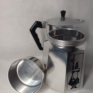 May include: A silver stovetop espresso maker, disassembled, with a black handle and a black knob on the lid. The base features a black graphic of a man with a hat. The filter basket is separate.