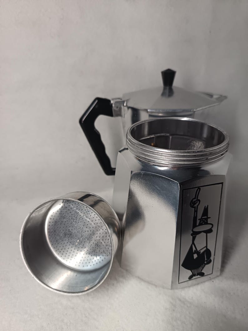 May include: A silver stovetop espresso maker, disassembled, with a black handle and a black knob on the lid. The base features a black graphic of a man with a hat. The filter basket is separate.