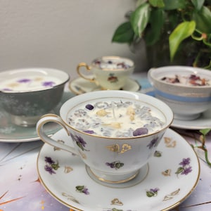 May include: A white teacup candle with a floral design and a purple and white wax blend. The candle is sitting on a white saucer with a floral design. There are other teacups and saucers in the background.