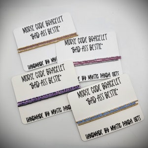 May include: Four handmade Morse code bracelets with the message "Bad Ass Bestie" on white cards. The bracelets are made with different colored beads, including gold, purple, pink, and blue.