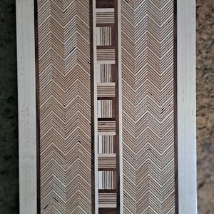May include: A rectangular wooden cutting board featuring a central vertical strip of alternating light and dark wood blocks. Flanking this center are two wide panels with a light and dark wood herringbone pattern, all enclosed by a light wood frame.