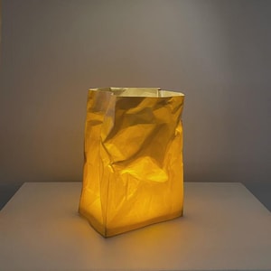 Puede incluir: Una lámpara rectangular con forma de bolsa de papel arrugada, que emite una cálida luz amarilla brillante desde su interior. La superficie exterior presenta un aspecto texturizado y plegado que difumina la iluminación, creando un efecto suave.