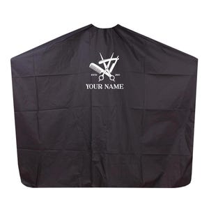 Puede incluir: Capa de peluquería negra con un gráfico blanco de tijeras, peine y navaja. El texto "YOUR NAME" está debajo del gráfico. La capa está diseñada para su uso en un salón de belleza.