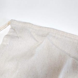 May include: A light beige drawstring pouch made of a woven fabric with a visible texture. The bag features a simple stitched hem at the top opening where a matching light beige cord is threaded through to allow for closure and cinching.