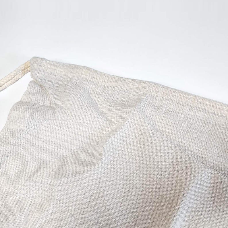May include: A light beige drawstring pouch made of a woven fabric with a visible texture. The bag features a simple stitched hem at the top opening where a matching light beige cord is threaded through to allow for closure and cinching.