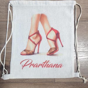 May include: A beige drawstring backpack featuring a printed illustration of two legs wearing red high-heeled sandals with crisscross straps. The name Prarthana is printed in red cursive script below the illustration of the footwear.