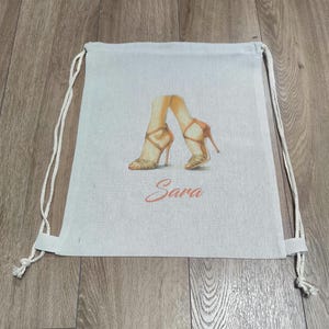 May include: A light beige drawstring backpack made of fabric, featuring a printed illustration of two legs wearing tan high-heeled sandals. Below the illustration, the name Sara is printed in a cursive, reddish-orange script font.