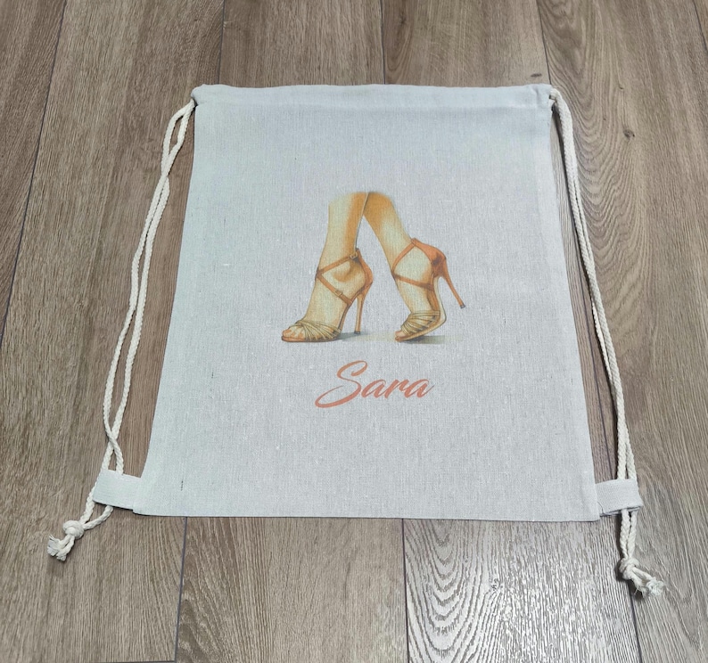 May include: A light beige drawstring backpack made of fabric, featuring a printed illustration of two legs wearing tan high-heeled sandals. Below the illustration, the name Sara is printed in a cursive, reddish-orange script font.