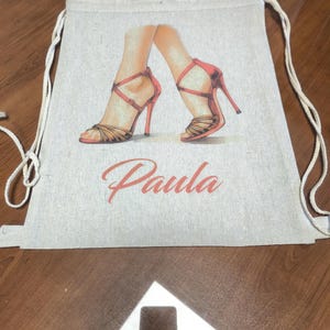 May include: A beige drawstring backpack made of a woven fabric featuring a printed illustration of two feet wearing red high-heeled sandals. The name Paula is printed in a cursive orange font below the illustration of the shoes.