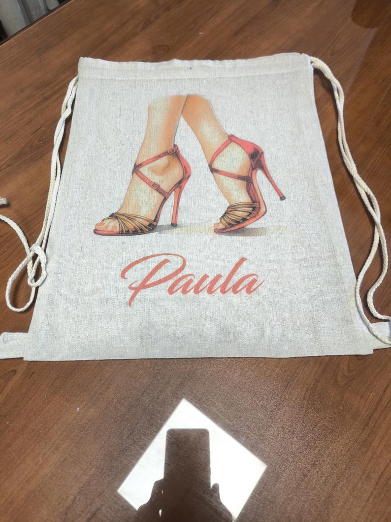 May include: A beige drawstring backpack made of a woven fabric featuring a printed illustration of two feet wearing red high-heeled sandals. The name Paula is printed in a cursive orange font below the illustration of the shoes.