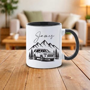 Peut inclure: Mug en céramique blanche avec intérieur et anse noirs. Le mug présente un graphisme noir et blanc d'une chaîne de montagnes, d'arbres et d'un véhicule. Le nom "James" est écrit en cursive au-dessus du graphisme.