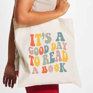 Może przedstawiać: Biała torba z płótna z napisem "It's a good day to read a book" w kolorowych, retro literach. Tekst jest otoczony małymi ilustracjami kwiatów.