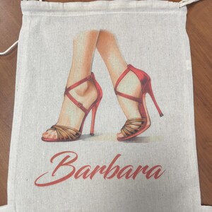 May include: A beige drawstring bag made of a woven fabric featuring a printed illustration of two legs wearing red high-heeled sandals with thin straps. The name Barbara is printed in a red cursive script font below the illustration.