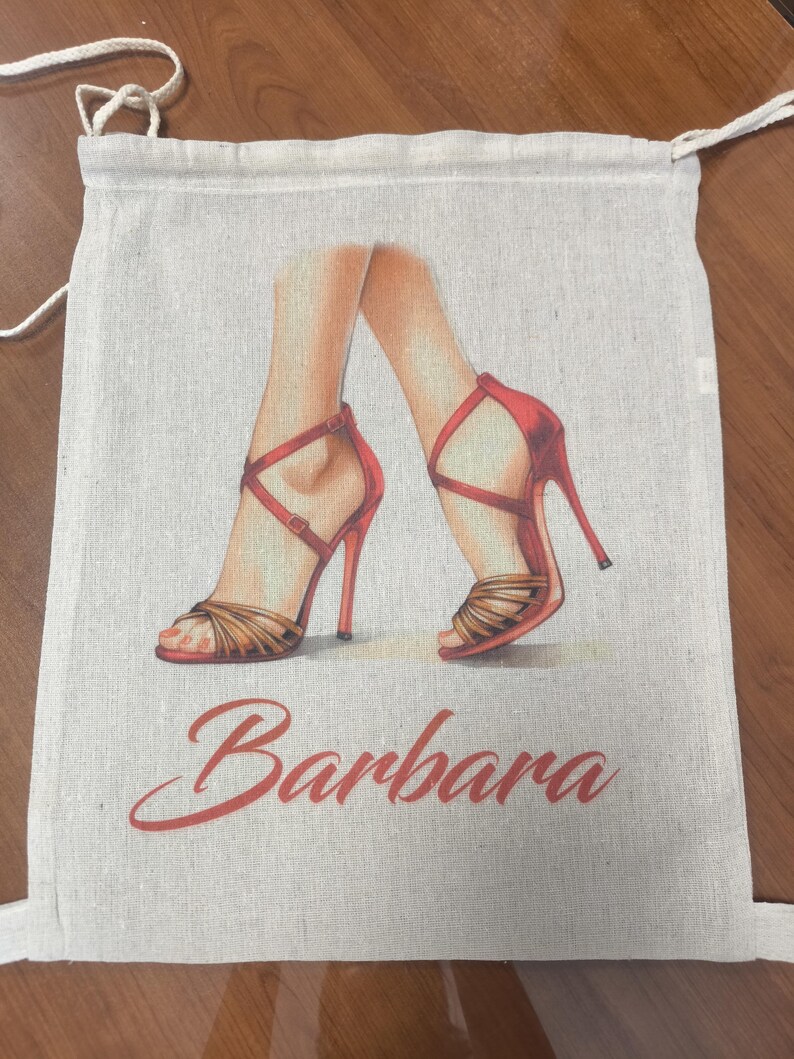 May include: A beige drawstring bag made of a woven fabric featuring a printed illustration of two legs wearing red high-heeled sandals with thin straps. The name Barbara is printed in a red cursive script font below the illustration.