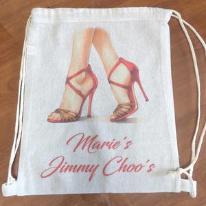 May include: A beige drawstring backpack made of canvas fabric featuring a printed illustration of legs wearing red high-heeled sandals with crisscross straps. The text Marie's Jimmy Choo's is printed in red cursive script below the illustration.