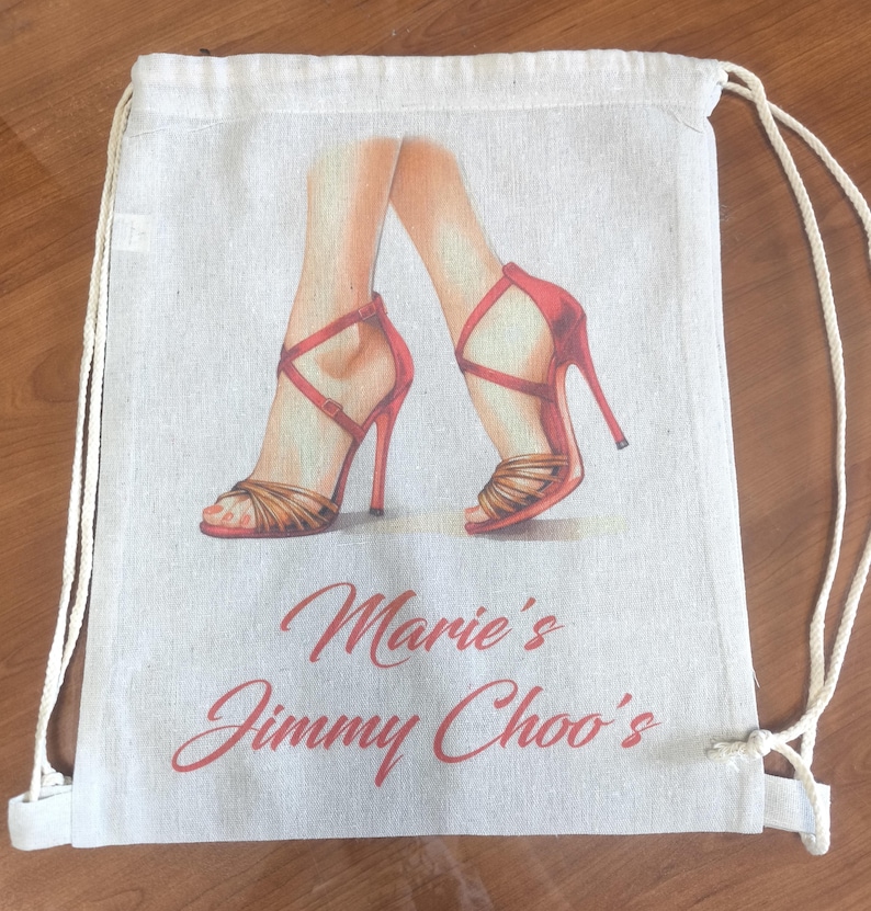 May include: A beige drawstring backpack made of canvas fabric featuring a printed illustration of legs wearing red high-heeled sandals with crisscross straps. The text Marie's Jimmy Choo's is printed in red cursive script below the illustration.