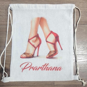 May include: A beige drawstring backpack featuring a printed illustration of two legs wearing red high-heeled sandals with crisscross straps. The name Prarthana is printed in a red cursive script font below the illustration of the footwear.