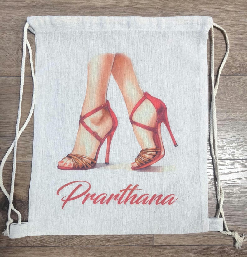 May include: A beige drawstring backpack featuring a printed illustration of two legs wearing red high-heeled sandals with crisscross straps. The name Prarthana is printed in a red cursive script font below the illustration of the footwear.