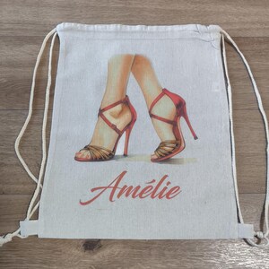 May include: A beige drawstring backpack made of canvas fabric. The front features a printed illustration of two legs wearing red high-heeled sandals with thin straps. Below the illustration, the name Am&eacute;lie is printed in red script.