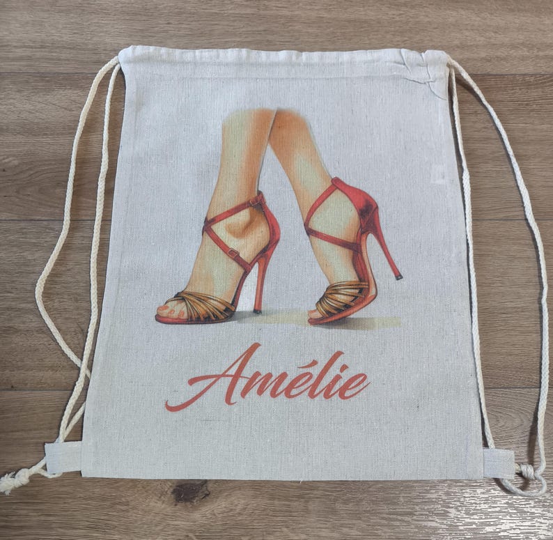 May include: A beige drawstring backpack made of canvas fabric. The front features a printed illustration of two legs wearing red high-heeled sandals with thin straps. Below the illustration, the name Am&eacute;lie is printed in red script.