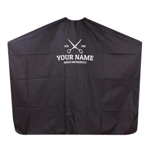 Puede incluir: Capa de barbero negra con texto blanco y un gráfico de tijeras. El texto dice "YOUR NAME" encima de "SHAVES AND HAIRCUTS" y "ESTD 1998". Esta capa protege la ropa durante cortes de pelo y afeitados.