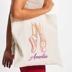Può includere: Una borsa tote bianca con un'illustrazione di ballerina rosa e il nome "Amelia" stampato in viola.