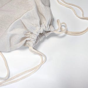 May include: A light beige drawstring bag made of a woven fabric with a visible texture. The bag features a gathered top closure secured by two matching beige braided cords that extend outward to form loops for carrying or hanging.