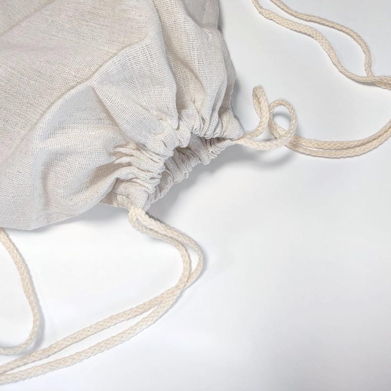 May include: A light beige drawstring bag made of a woven fabric with a visible texture. The bag features a gathered top closure secured by two matching beige braided cords that extend outward to form loops for carrying or hanging.