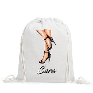 May include: A white drawstring backpack featuring a graphic illustration of two legs wearing black high-heeled sandals. The name Sara is printed in a black cursive script font below the illustration of the legs on the front of the bag.
