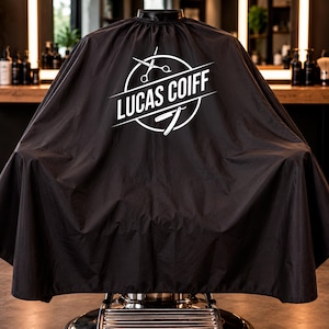 Könnte beinhalten: Ein schwarzer Friseurumhang mit einem weißen kreisförmigen Logo, das den Text LUCAS COIFF sowie grafische Darstellungen einer Schere und eines Rasiermessers zeigt. Der Umhang hat einen hohen Kragen und fällt in einer weiten Silhouette.