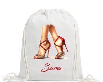 Puede incluir: Bolsa con cordón blanca con una ilustración de piernas con zapatos de tacón alto rojos y dorados. El nombre "Sara" está escrito en rojo debajo. La bolsa es de tela clara.