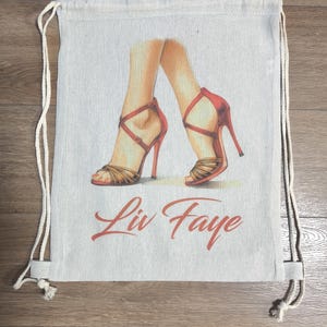 May include: A beige drawstring backpack featuring a graphic illustration of two legs wearing red high-heeled strappy sandals. Below the illustration, the text Liv Faye is printed in a red cursive font. The bag has white rope straps.