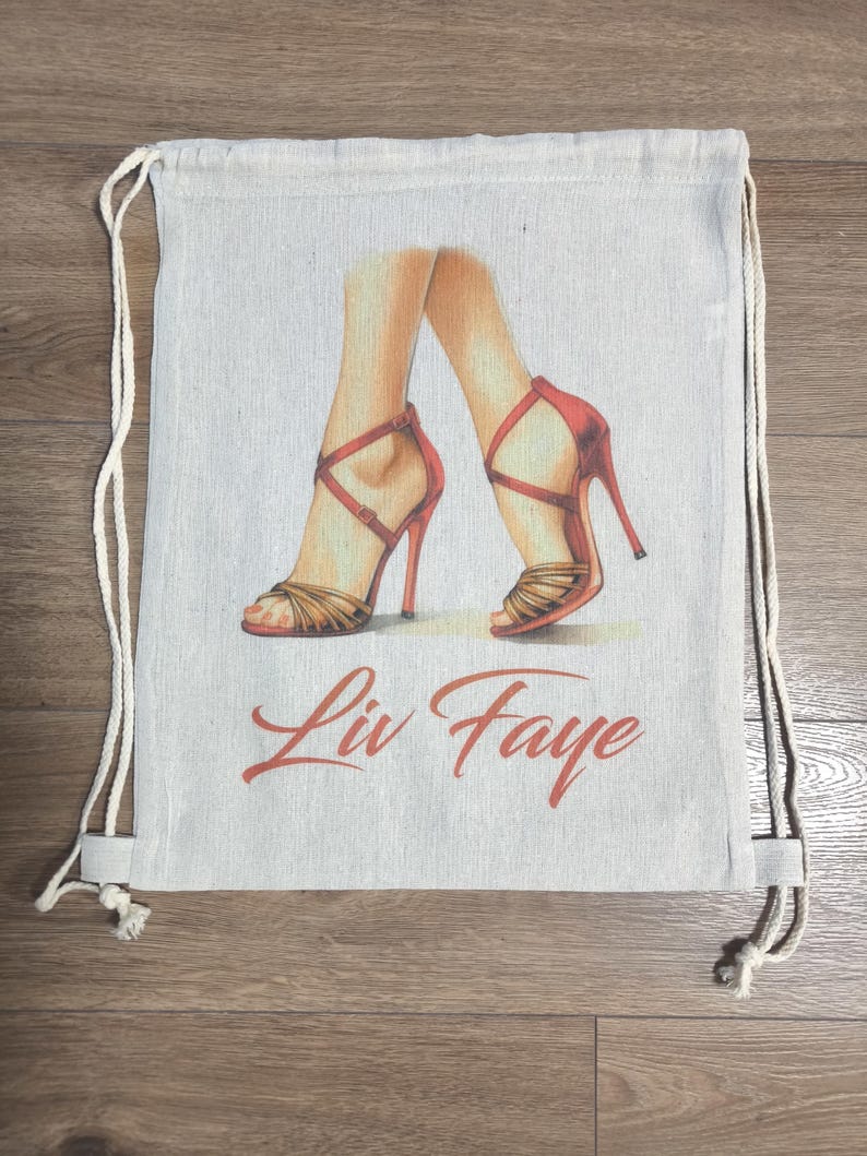May include: A beige drawstring backpack featuring a graphic illustration of two legs wearing red high-heeled strappy sandals. Below the illustration, the text Liv Faye is printed in a red cursive font. The bag has white rope straps.