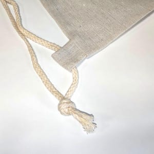 May include: A light beige drawstring bag made of a woven fabric with a visible texture. The bag features a thick, braided off-white cord that passes through a fabric loop at the top corner and is secured with a simple knot at the end.