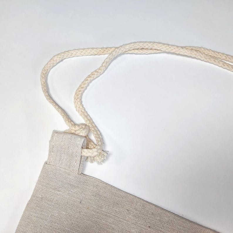 May include: A light beige drawstring bag made of a woven fabric texture. The bag features thick, braided off-white cotton rope handles that are threaded through a fabric loop at the top corner, creating a simple and functional design.
