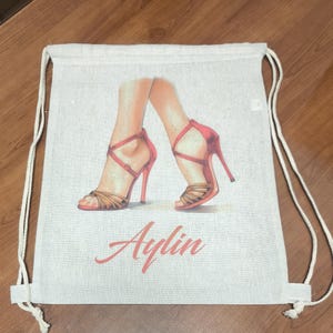 May include: A beige drawstring backpack featuring a printed illustration of two legs wearing red high-heeled sandals with thin straps. The name Aylin is printed in a cursive red font below the illustration of the legs and shoes.