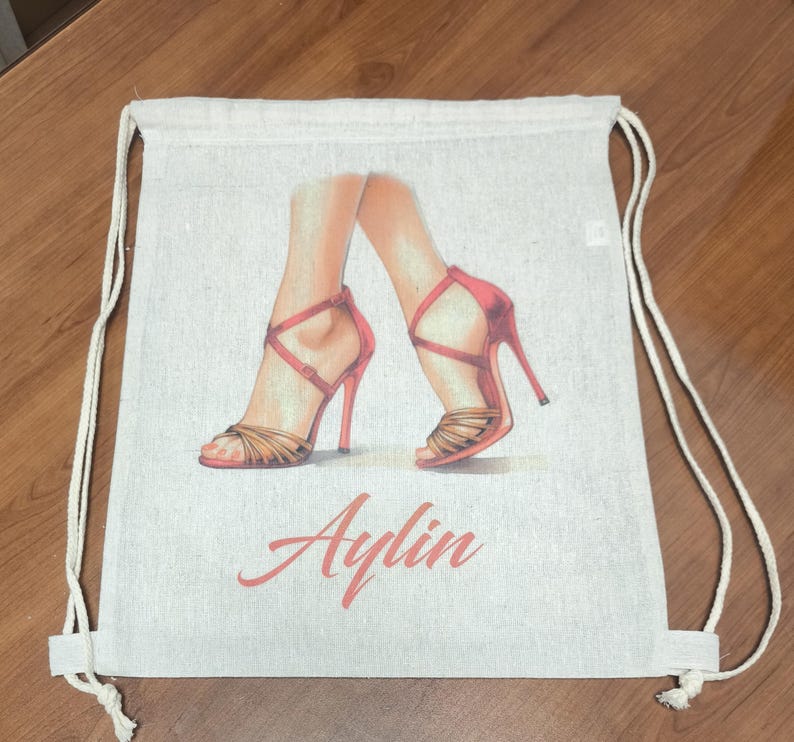 May include: A beige drawstring backpack featuring a printed illustration of two legs wearing red high-heeled sandals with thin straps. The name Aylin is printed in a cursive red font below the illustration of the legs and shoes.