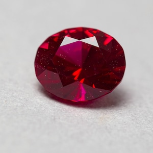 Pode incluir: Uma gema de rubi vermelho-escuro com corte facetado. A gema tem formato oval e alto polimento.