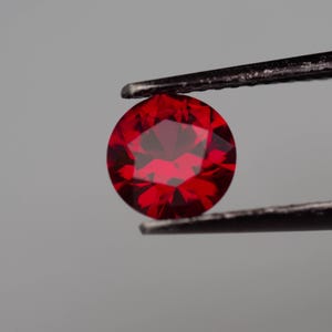 Pode incluir: Uma gema vermelha profunda com corte facetado, segurada por pinças contra um fundo cinza.