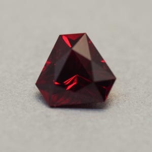 May include: A faceted, deep red gemstone with a unique geometric shape. The gem has multiple facets that reflect light, creating a brilliant sparkle. The stone is set against a neutral grey background.