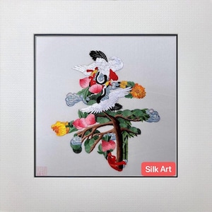 May include: Embroidered silk art featuring a tree with white cranes, pink and yellow flowers, and green foliage. The artwork is framed in a white mat and black frame. The text "Silk Art" is in a red rectangle.