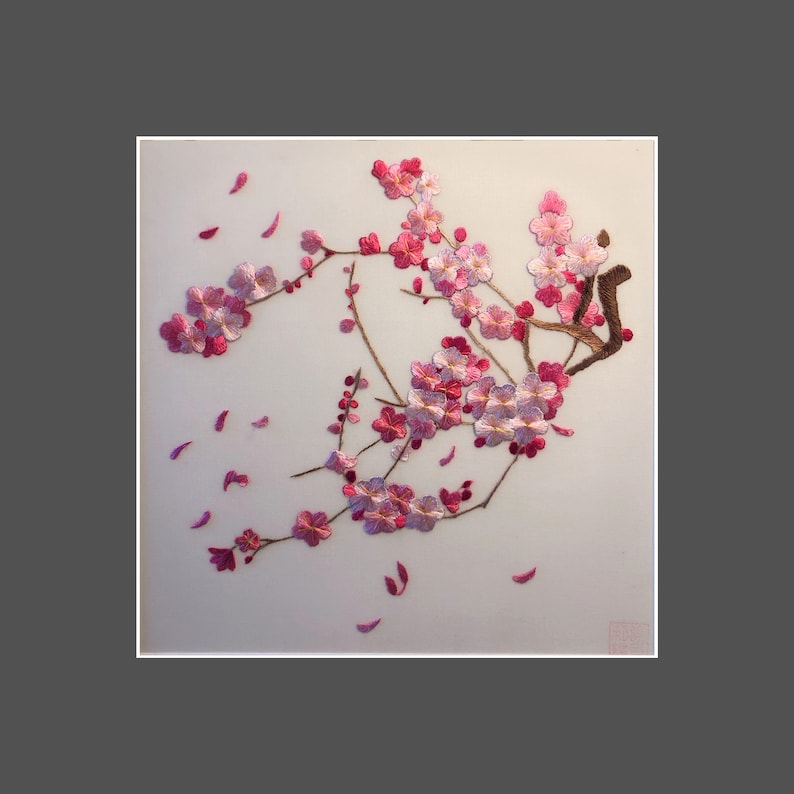 May include: Embroidered artwork featuring a branch of pink cherry blossoms with falling petals. The blossoms are detailed with varying shades of pink and white, set against a light background. The artwork is framed in white.