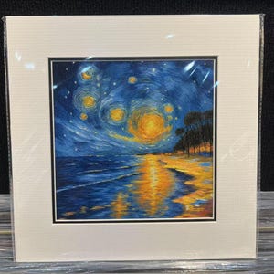 May include: A framed art print depicting a night scene with a swirling, starry sky in shades of blue and yellow. The artwork shows a beach with the reflection of the sky in the water. The print is matted and framed in a white border.